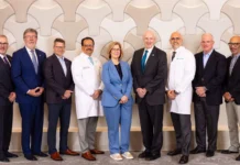 Sutter Health and Allina Health Sign Definitive Agreement to Join Forces Group of nine healthcare professionals standing in a line, some in white coats and others in suits, posing for a team photo in front of a decorative wall.