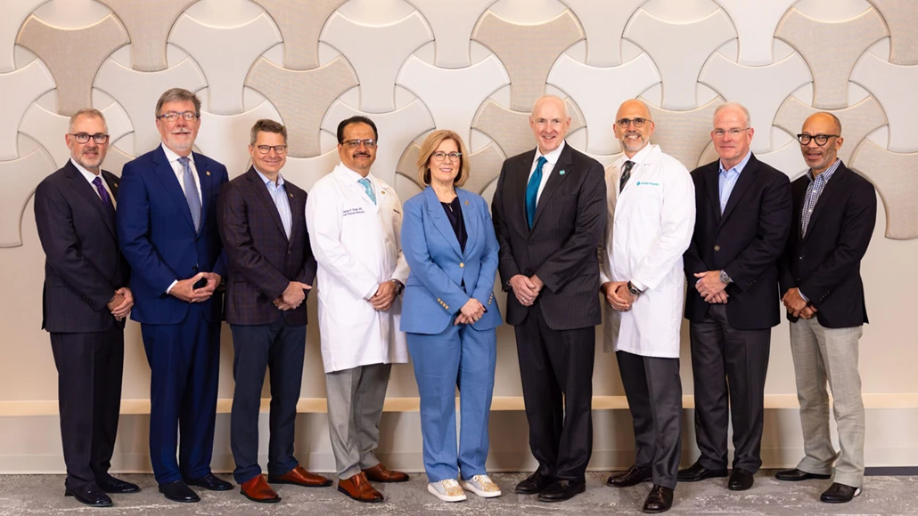 Group of nine healthcare professionals standing in a line, some in white coats and others in suits, posing for a team photo in front of a decorative wall.