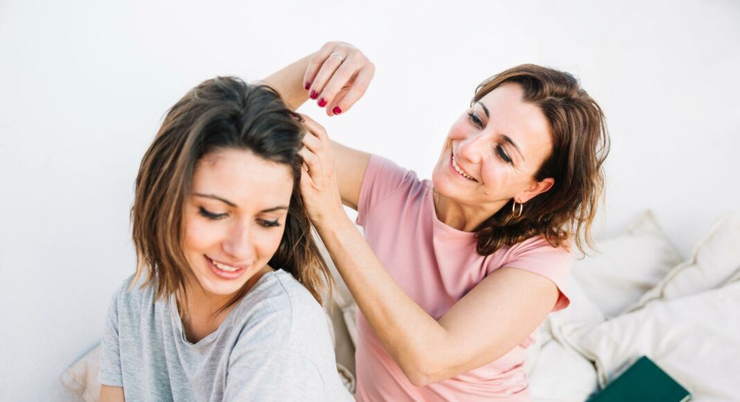 Two women sit together on a sofa; one braids the other's hair and both smile.