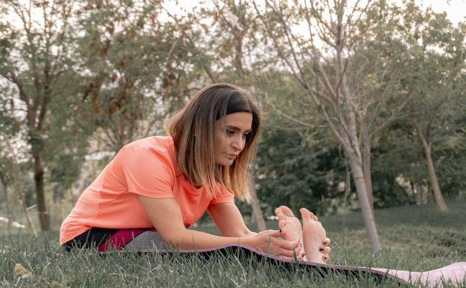 Woman in a coral T-shirt sitting on a yoga mat in a park, bending forward to touch her toes while stretching outdoors.