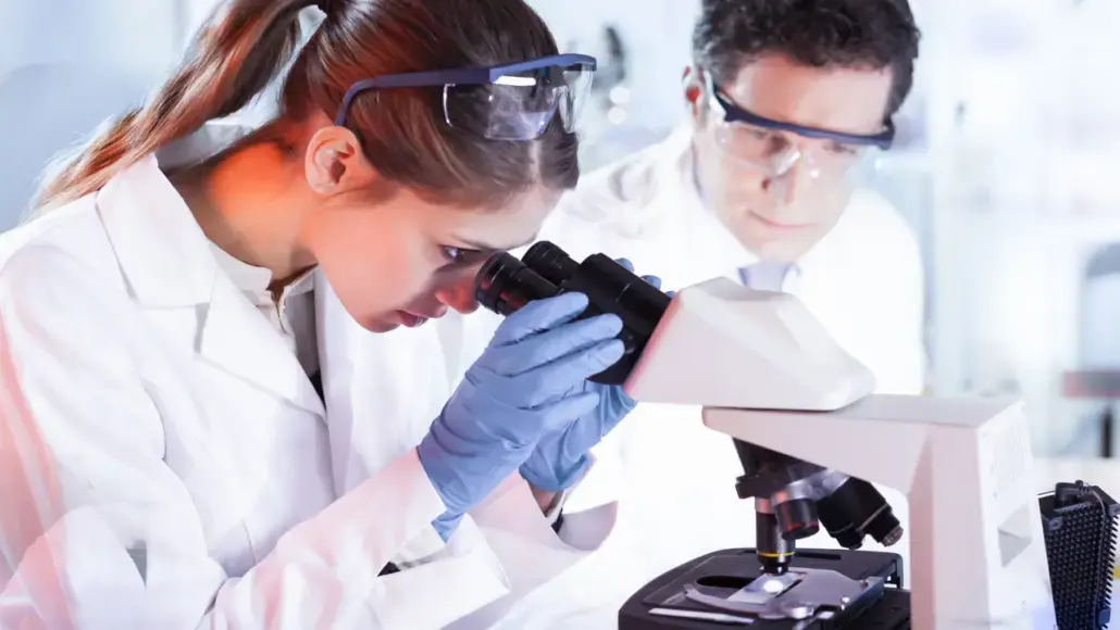 Female scientist examines slides through a compound microscope, wearing gloves and safety goggles, with a male colleague in the background.