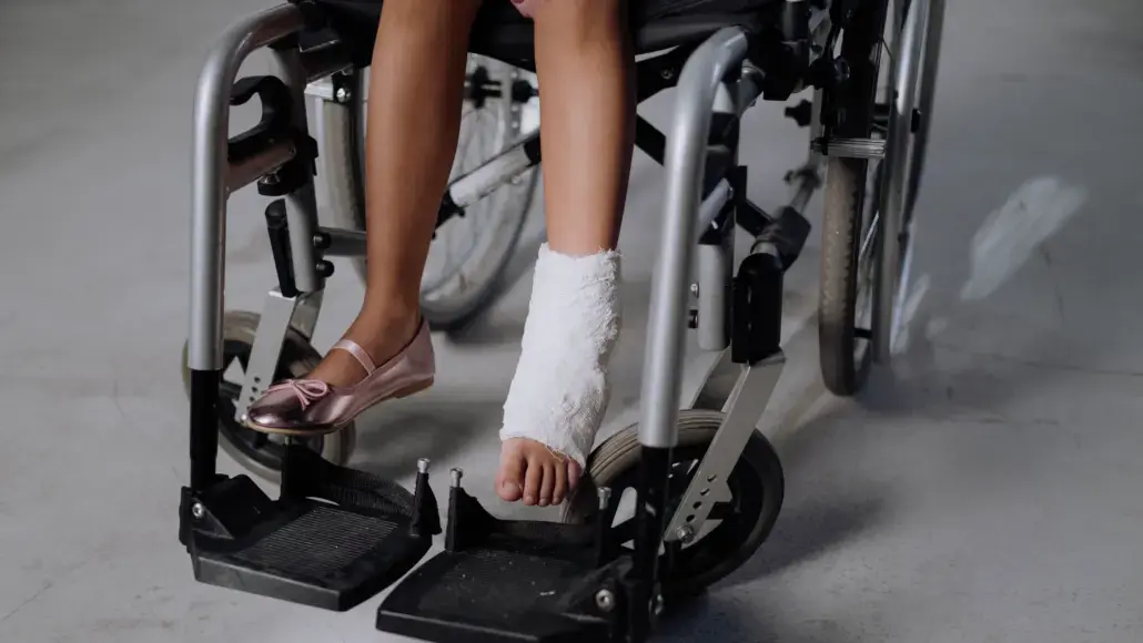 Person seated in a wheelchair with a white bandaged leg cast extending from the ankle to mid-calf, and a pink flat shoe on the other foot above the footrest.