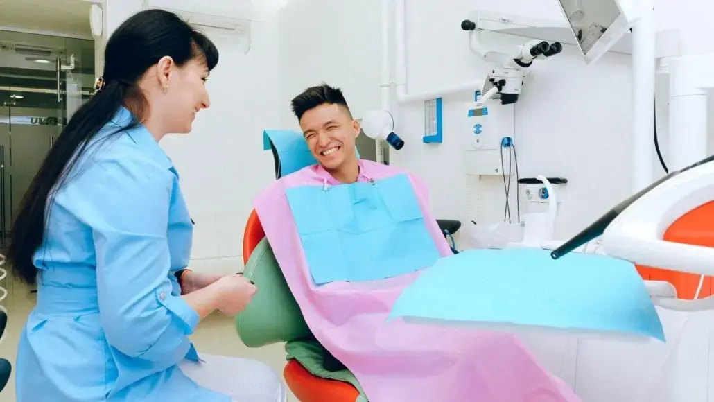 A patient in a dental chair smiles at a dental professional seated beside him in a bright clinic, both wearing bibs and masks nearby (informational).