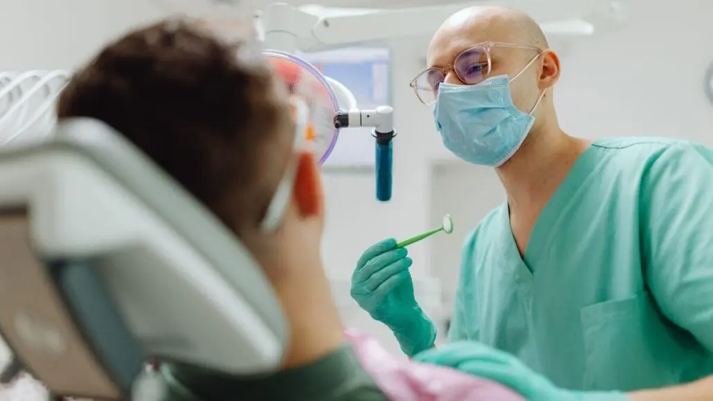 Dentist in scrubs, mask, and gloves examines a patient in a dental chair, holding a dental tool under bright clinic lighting.